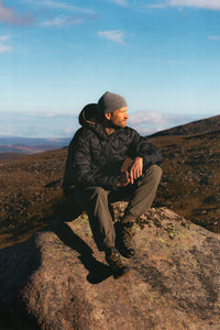 A man sat on a rock, wearing the KESTIN Cuillin Down Jacket in Black.