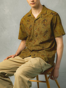 A man sitting on a chair, wearing a green floral short sleeve shirt.